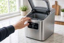 A hand reaches toward an open-top stainless steel ice maker on a kitchen counter, ready to start ice production.