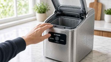 A hand reaches toward an open-top stainless steel ice maker on a kitchen counter, ready to start ice production.